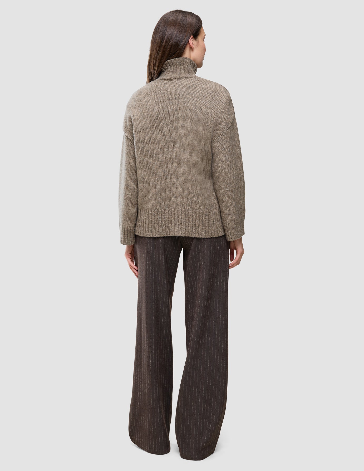 Eine Person, von hinten gesehen, die einen taupefarbenen, melierten Damen-Rollkragenpullover aus dicker, weicher Wolle und eine dunkle Nadelstreifenhose trägt.